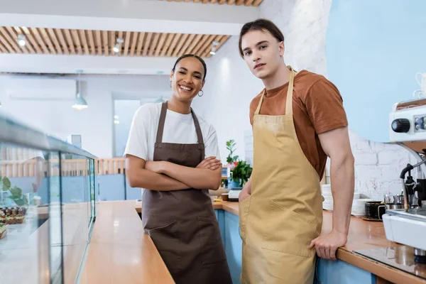 Cheerful multiethnic sellers looking at camera near showcase and coffee machine in sweet shop — Stock Photo