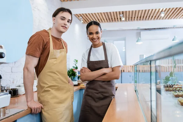 Smiling multiethnic sellers posing near showcase in confectionery - foto de stock