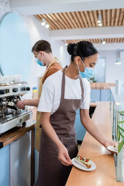 African american seller in medical mask taking dessert from showcase near colleague in confectionery — стокове фото