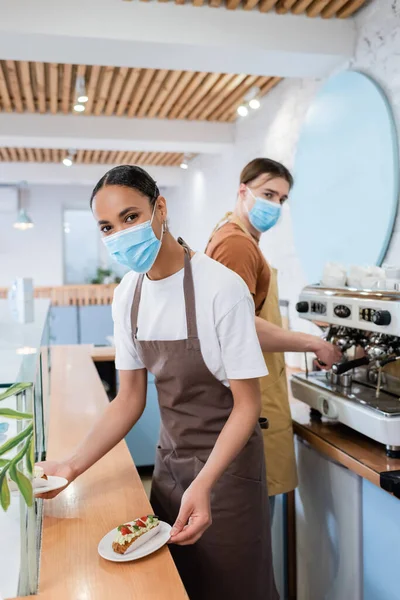 African american seller in medical mask holding eclairs and looking at camera near blurred barista in sweet shop - foto de stock