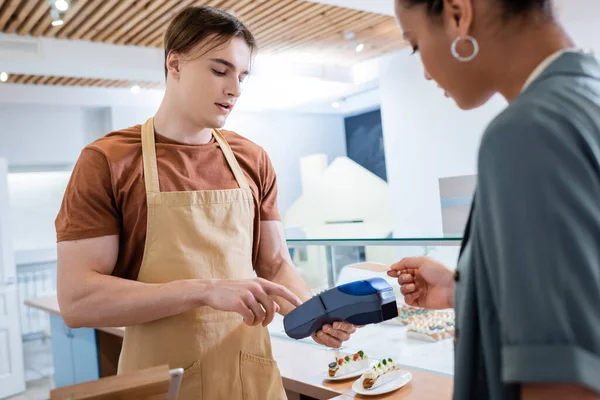 Verkäufer hält Zahlungsterminal in Nähe verschwommener afrikanisch-amerikanischer Kundin in Süßwarenladen — Stockfoto