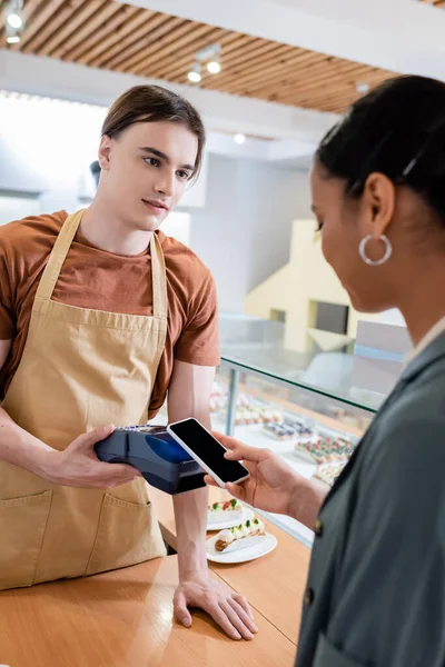 Young salesman holding payment terminal near african american customer with smartphone in confectionery — стоковое фото