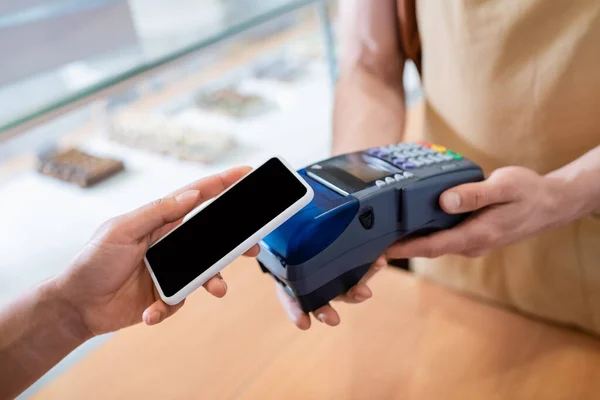 Cropped view of african american woman paying with smartphone near salesman in confectionery — стокове фото