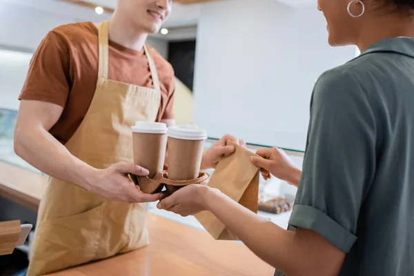 Cropped view of african american customer taking coffee to go and paper bag from seller in confectionery — Foto stock