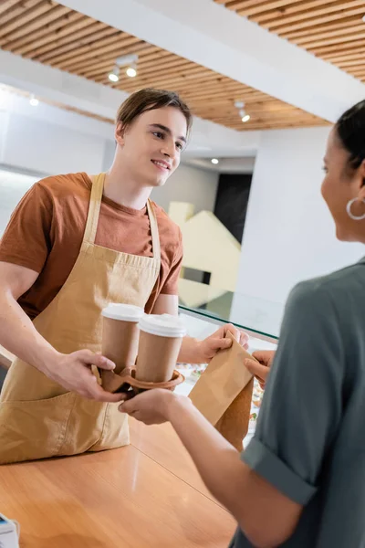 Lächelnder Verkäufer gibt Kaffee to go und Papiertüte an verschwommenen afrikanisch-amerikanischen Kunden in der Konditorei — Stockfoto