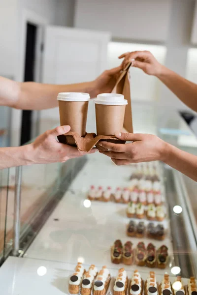 Cropped view of salesman giving paper bag and cups to african american client near showcase in confectionery — стоковое фото