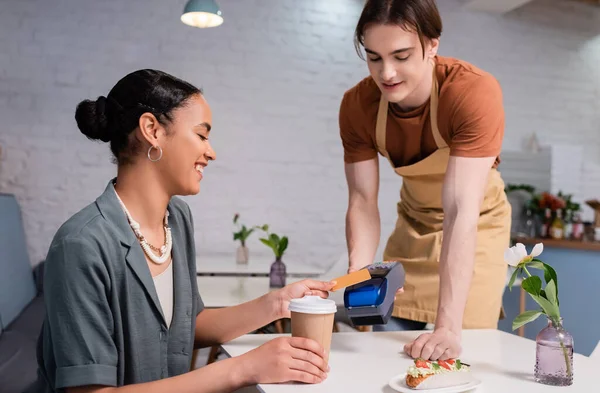 Positive african american customer paying with credit card near salesman and dessert in confectionery — Foto stock