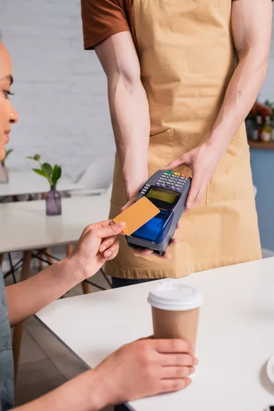 Salesman holding payment terminal near african american client with coffee in sweet shop — стоковое фото