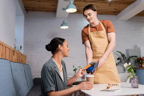 Lächelnder afrikanisch-amerikanischer Kunde bezahlt mit Kreditkarte in der Nähe von Verkäufer und Eclair im Süßwarenladen — Stockfoto