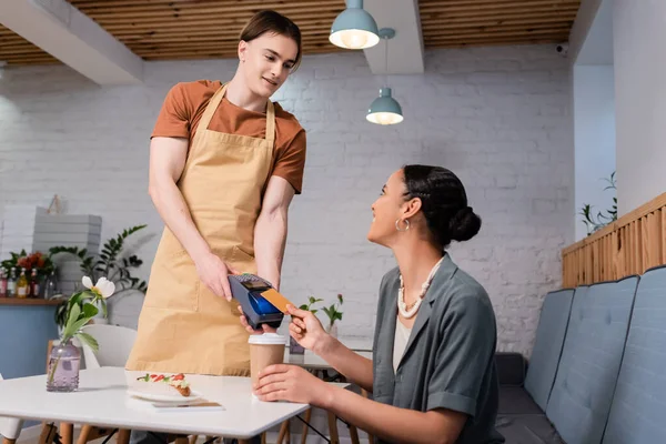 Smiling seller holding payment terminal near african american customer with coffee in confectionery — Stock Photo