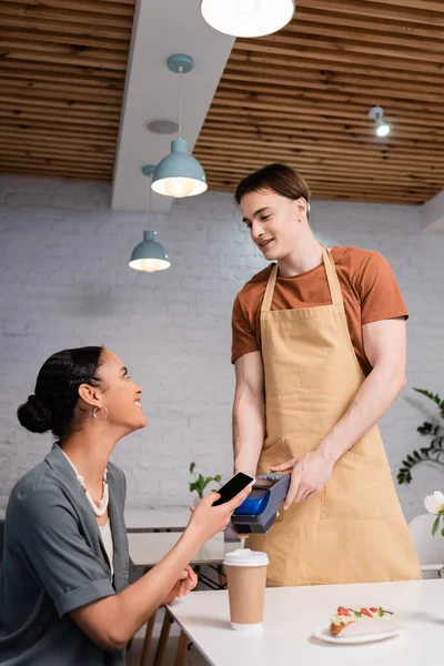 Positive african american woman paying with smartphone near salesman and dessert in confectionery — стокове фото