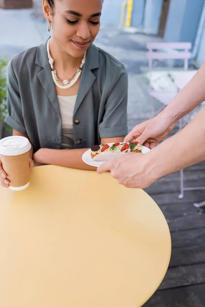 Salesman holding eclair near african american woman with coffee to go on terrace of confectionery — Fotografia de Stock