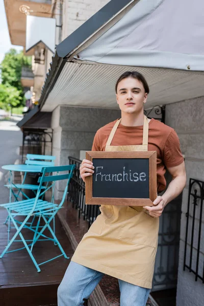 Young salesman holding chalkboard with franchise lettering on terrace of confectionery — Stock Photo
