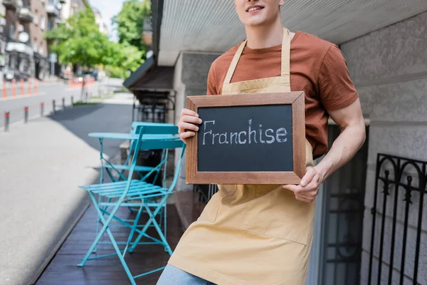 Cropped view of smiling seller holding chalkboard with franchise lettering near sweet shop on urban street — Photo de stock