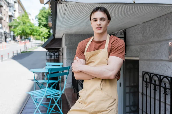 Young salesman in apron crossing arms on terrace of confectionery — Photo de stock