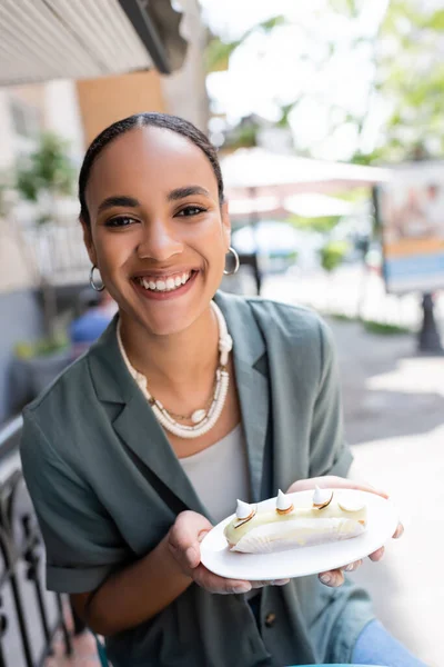 Позитивний афроамериканець тримає десерт і дивиться на фотоапарат на терасі солодкої крамниці. — стокове фото