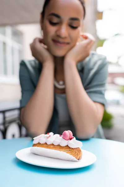 Tasty eclair near blurred african american woman on terrace of sweet shop — Stockfoto
