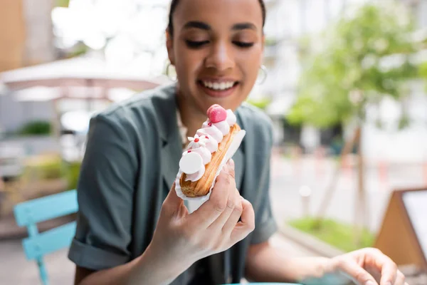 Verschwommen lächelnde afrikanisch-amerikanische Kundin hält Eclair auf der Terrasse eines Süßwarenladens — Stockfoto