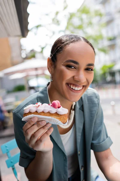 Positive afrikanisch-amerikanische Kundin hält Eclair mit Sahne auf der Süßwarenterrasse — Stockfoto