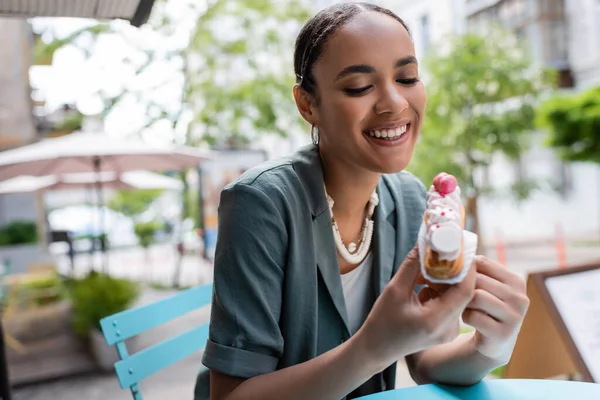 Pretty african american woman looking at eclair with cream on terrace of confectionery — Photo de stock