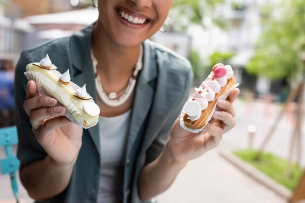 Обрізаний вид усміхненого афроамериканського клієнта, який тримає смачні еклери на терасі кондитерських виробів — стокове фото