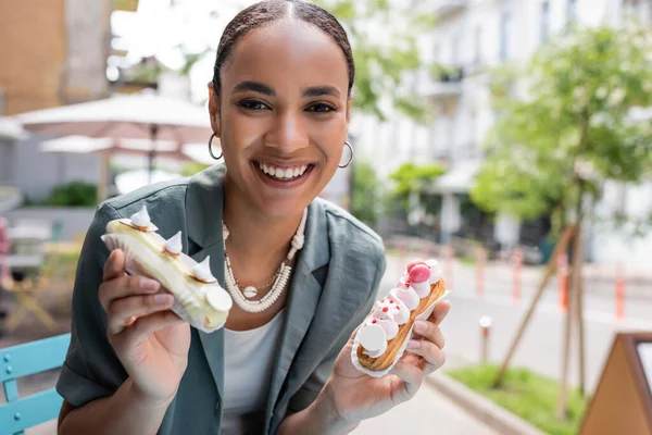 Positive african american woman holding delicious eclairs on terrace of confectionery — стоковое фото