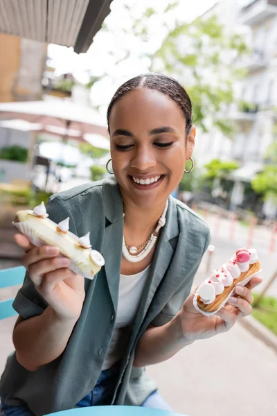 Smiling african american woman looking at eclairs with cream on terrace of confectionery — стокове фото