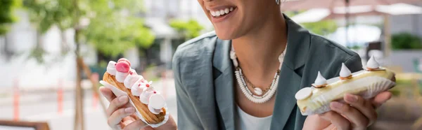 Обрізаний вид веселого афроамериканського клієнта, який тримає еклери на терасі солодкого магазину, банер — стокове фото