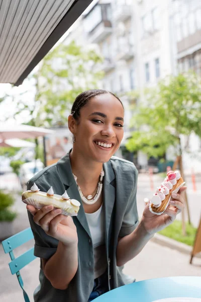 Smiling african american client holding eclairs and looking at camera on terrace of sweet shop - foto de stock