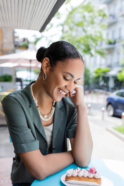 Cheerful african american woman looking at delicious eclair on terrace of sweet shop — стоковое фото