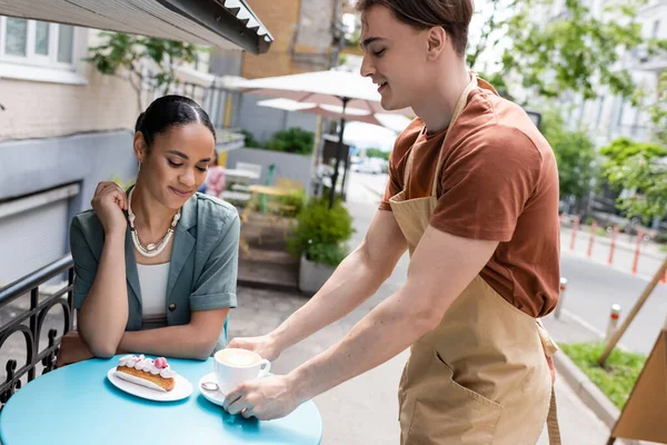 Smiling seller holding cup of coffee near dessert and african american customer on terrace of sweet shop - foto de stock