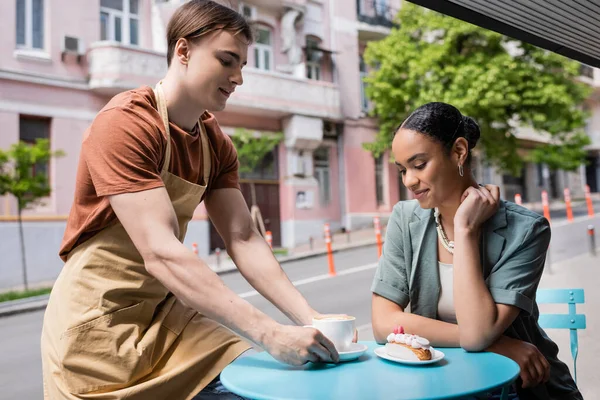 Seller in apron putting cappuccino near dessert and african american client on terrace of sweet shop — стоковое фото