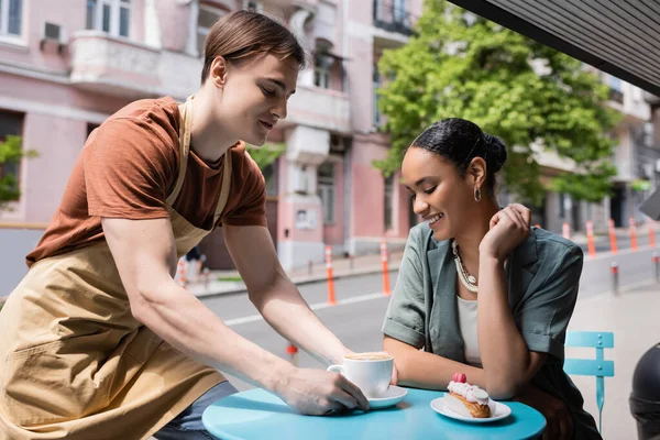 Smiling african american customer sitting near dessert and seller with coffee on terrace of confectionery — Stockfoto