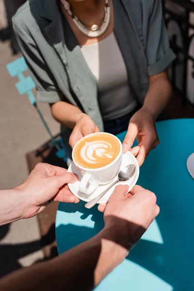 Cropped view of waiter giving cup of cappuccino to african american client on terrace of sweet shop — Stockfoto