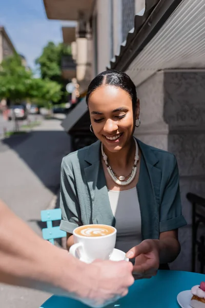 Smiling african american woman taking cappuccino from waiter near dessert on terrace of sweet shop - foto de stock