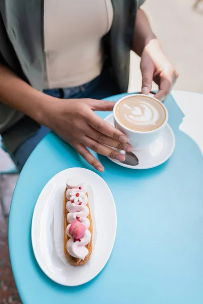 High angle view of delicious eclair near blurred african american woman with cup of cappuccino on terrace of confectionery — Stock Photo