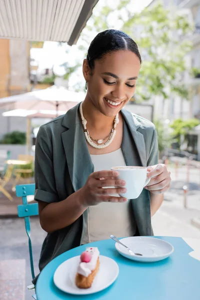 Positive african american woman holding cappuccino near blurred dessert on terrace of sweet shop - foto de stock