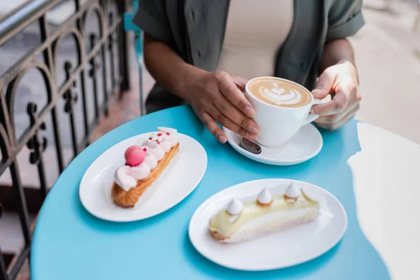 Ausgeschnittene Ansicht einer afrikanisch-amerikanischen Frau mit einer Tasse Cappuccino in der Nähe von Eclairs auf der Terrasse eines Süßwarengeschäfts — Stockfoto