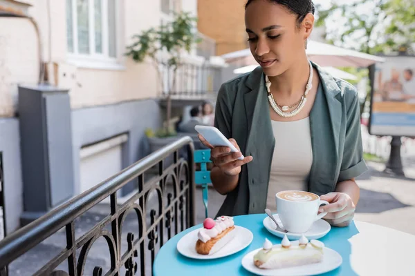 Afroamerikanerin benutzt Smartphone in der Nähe von Kaffee und Eclairs auf der Terrasse eines Süßwarengeschäfts — Stockfoto