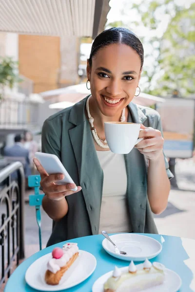 Smiling african american client holding coffee and smartphone near eclairs on terrace of sweet shop — Photo de stock