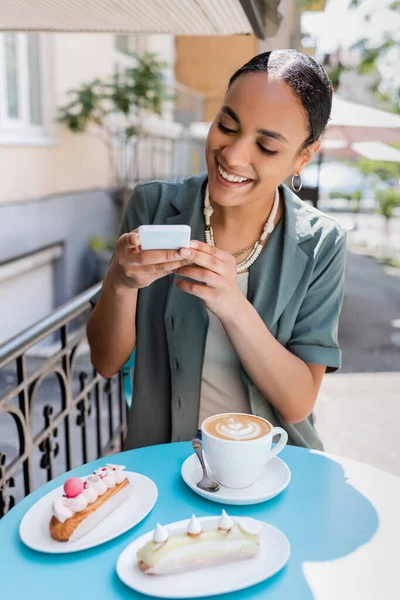 Smiling african american client taking photo on smartphone near coffee and dessert on terrace of sweet shop - foto de stock
