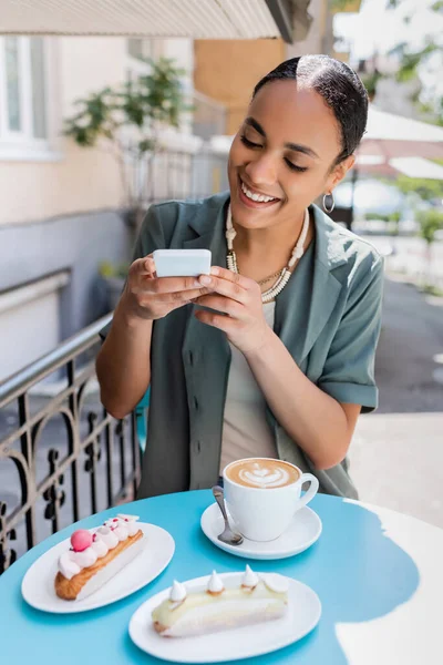 Smiling african american client taking photo on smartphone near coffee and dessert on terrace of sweet shop — стоковое фото