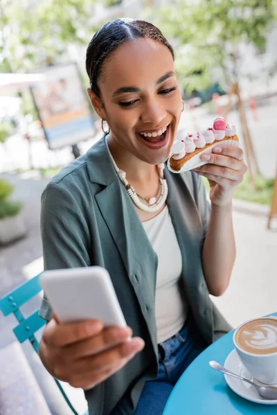 Lächelnder afrikanisch-amerikanischer Kunde hält Eclair in der Hand und macht ein Selfie auf dem Smartphone in der Nähe von Kaffee auf der Terrasse eines Süßwarengeschäfts — Stockfoto
