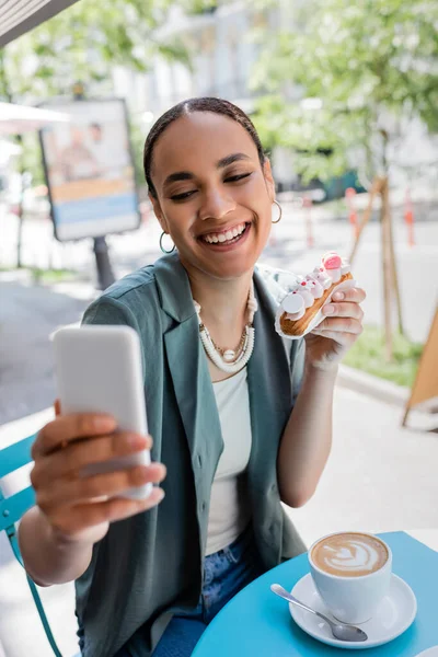 Smiling african american woman holding eclair and taking selfie on smartphone near cappuccino on terrace of sweet shop — стоковое фото