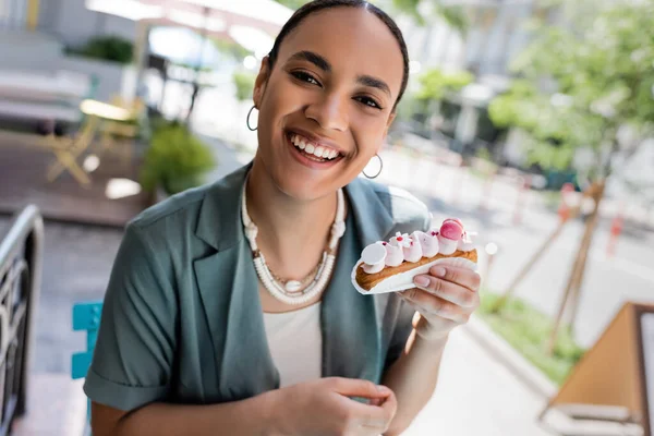 Cheerful african american woman holding eclair and looking at camera on terrace of cafe — Fotografia de Stock