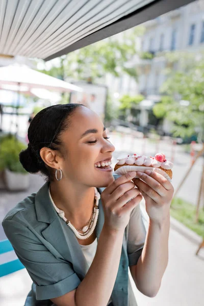 Young african american woman eating tasty eclair on terrace of sweet shop — Foto stock