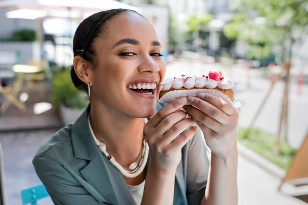Cheerful african american woman holding eclair and looking at camera outdoors — стоковое фото