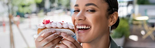 Happy african american woman holding delicious eclair on terrace of sweet shop, banner — стокове фото