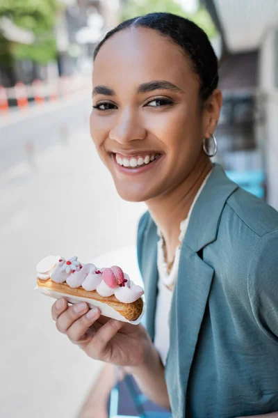 Pretty young customer holding delicious eclair and looking at camera on terrace of cafe - foto de stock