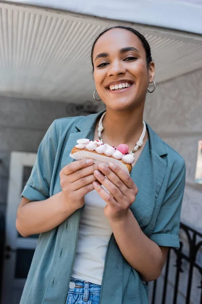 Усміхнена афроамериканка тримає смачний еклер біля солодкого магазину — стокове фото